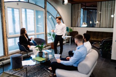 Zarif kıyafetli genç lider modern mobilya ve pencereli konferans salonunda iş arkadaşlarıyla iş görüşmesi yapıyor.