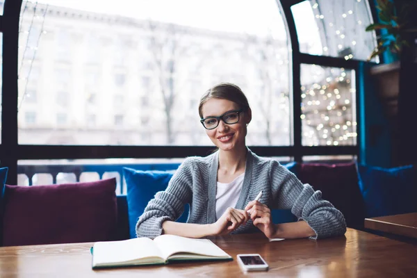 Young Cheerful Female Gathered Hair Glasses Looking Camera While Sitting — стоковое фото