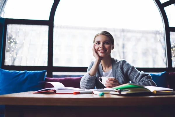 休憩中にノートやコーヒーカップとテーブルに座っている間に離れて見て頬に歯の笑顔と手で楽観的な女性のフリーランサー — ストック写真