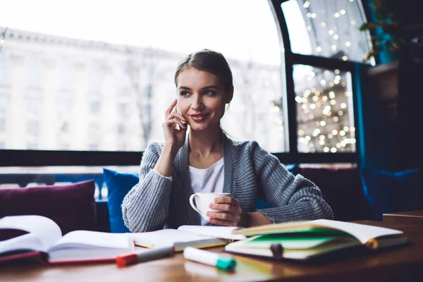 若い幸せな女性のリモートワーカー離れてコーヒーを飲みながらテーブルに座って仕事で休憩をしながら電話をかける — ストック写真