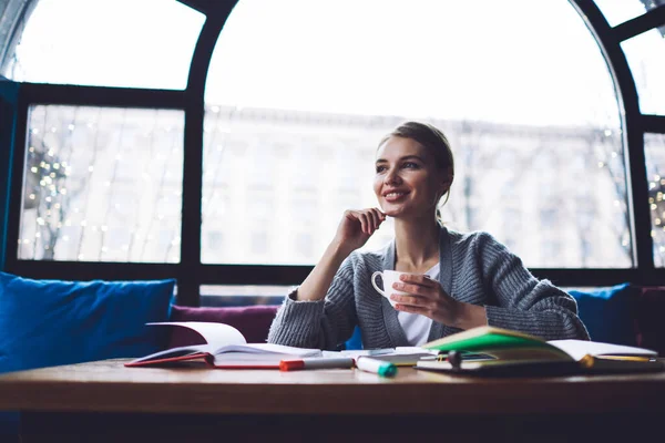休憩中に教科書やコーヒーカップとテーブルに座っている間に離れて見てあごで手でカジュアルな服を着た若い幸せな女性学生 — ストック写真