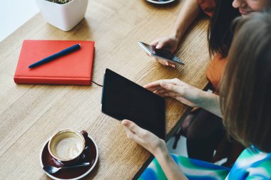 Yukarıdaki arkadaşlardan, elinde kahve ve kırmızı defterle masada oturan akıllı telefonu kullanarak tablete fotoğraf yükleyen.