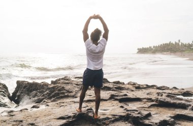 Kumlu sahilde kollarını kaldırmış ve deniz manzarasına hayran kalmış, sıradan giysili, tanınmayan bir adamın arka görünüşü.