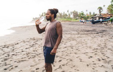 Plastik şişeden su içerken bulutlu bir yaz gününde okyanusun kıyısındaki kumlu tropikal sahilde duran sakallı siyah erkeğin yan görüntüsü.