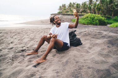 Yaz günü kumlu sahil şeridinde dinlenirken günlük kıyafetleriyle gülümseyen ve cep telefonuyla selfie çeken genç Afrikalı Amerikalı erkek gezgin.