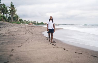 Siyah, uzun boylu, günlük giysiler içinde sahilde yürüyen ve bulutlu bir günde kameraya gülümseyen bir adam.