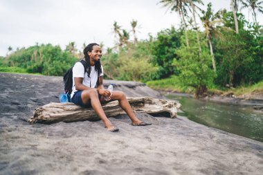 Çıplak ayaklı, Afro-Amerikalı, sıradan kıyafetli, egzotik bir şekilde ağaç gövdesinde dinlenirken gülümseyen ve yüzünü çeviren bir erkek.