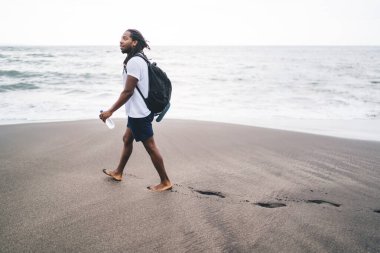 Sırt çantasında su şişesi taşıyan ve kıyı şeridinde gezinen Afro-Amerikan erkek.