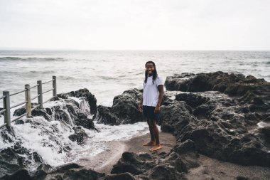 Tüm vücudun yüksek açılı mutlu Afrikalı Amerikalı erkek kayalık sahilde durup gülümserken kameraya bakıyor.
