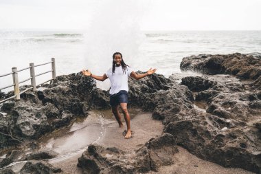 Tüm vücudun yüksek açılı pozitifti. Sıradan giyinmiş, gülümseyen ve kollarını kayalık sahilde açmış kameraya bakan Afro-Amerikan erkek.