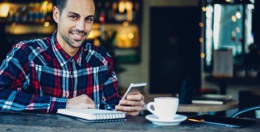 Hafta sonu kahve dükkanında kameraya gülümseyen internet haberlerini kontrol etmek için planlamacı defteri ve modern akıllı telefon cihazıyla mutlu müşterilerin yarım boy portresi, cep telefonlu neşeli blogcu