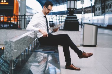 Gözlük takan, gözlük takan, finans gazetesi okuyan, Kafkas erkek girişimci, platformun yakınında demiryolu bekleyen kitle iletişim araçlarından bilgi kontrol eden kendine güvenen işadamının yan görüntüsü