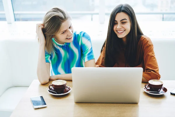从上图中我们可以看到 年轻女性面带微笑地坐在桌旁 一边拿着智能手机和咖啡 一边在笔记本电脑上学习 — 图库照片