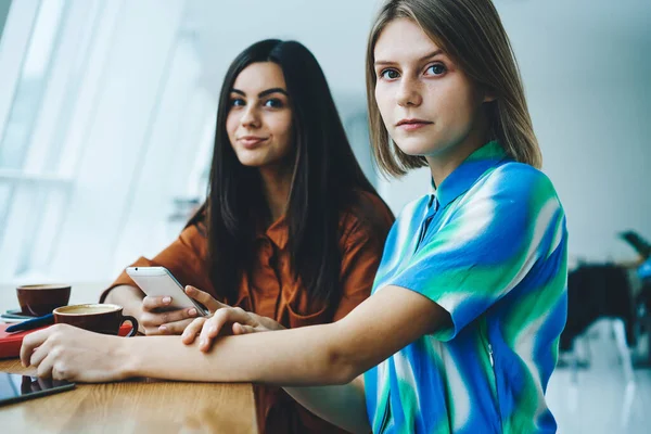 Genç bayan arkadaşlar, kafeteryada gündüz vakti kahve ve akıllı telefonlar eşliğinde günlük kıyafetlerini giyerken kameraya bakarlar.