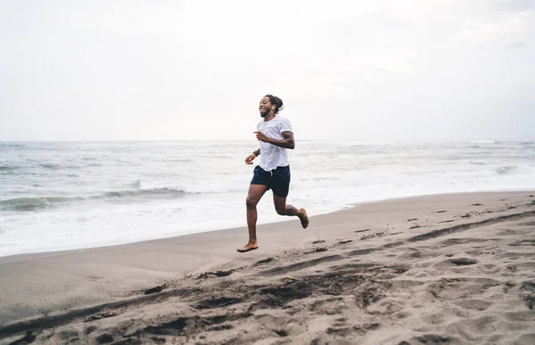 全身裸足アフリカ系アメリカ人男性ジョガーでスポーツウェアで実行中砂のビーチ近くに波海と朝のイヤホン経由で音楽を聞く — ストック写真