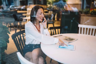 Havalı giyinmiş, gülümseyen olgun bir bayandan akıllı telefondan konuşmak ve modern kafenin penceresinde otururken tablet üzerinde sörf yapmak.