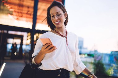 Mobil uygulama üzerinden seyahat yayınını paylaşmak için 4G 'ye bağlanan mutlu beyaz blog yazarı, akıllı telefon teknolojisiyle sohbet ederken gülümseyen klasik gözlüklü neşeli kadın