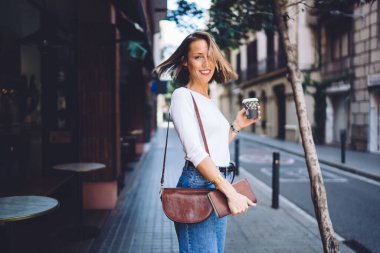 Gündelik kıyafetlerle gülümseyen beyaz kadının yarım boy portresi aromalı kahvenin tadını çıkarıyor ve tatillerde şehir merkezinde boş zamanlarının tadını çıkarıyor. Kahve içen güzel bir kadın boş zamanlarında kameraya bakıyor.