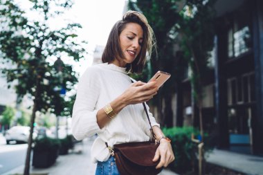 Yeşil şehir caddesinin bulanık arka planında dikilirken şık bir bayanın akıllı telefonuyla mesajlaşması.