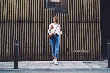 Moda kıyafetleri içinde uzun boylu neşeli bir bayan akıllı telefonda sohbet ediyor ve şehir sokaklarında vakit geçirirken kahve içiyor.