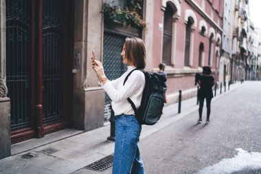 Kafkasyalı turistin günlük kıyafetleriyle gezerken cep kamerasıyla binaları çekerken yan görüntüsü, mobil cihazlı kız sosyal ağlara paylaşmak için fotoğraf çekiyor.