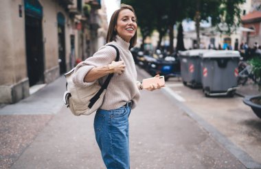 Gezici sırt çantalı neşeli bayan turist şehir sokağında duruyor ve tek başına tatile çıkmaktan memnun, modern mobil teknolojisi olan mutlu beyaz kadın eğleniyor.