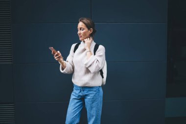 Sırt çantalı, mavi duvarın yanında duran ve ekrana bakarken cep telefonunu karıştıran normal giyinmiş yetişkin bir kadın.