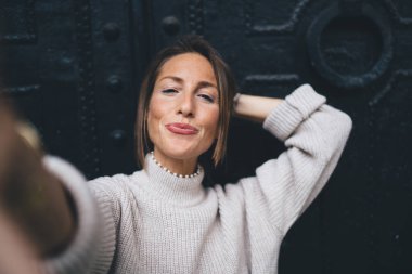 Neşeli bir kadın günlük giysiler içinde saçlarını tek eliyle temizliyor ve akıllı telefondan selfie çekiyor ve demir kapıya bakıyor.