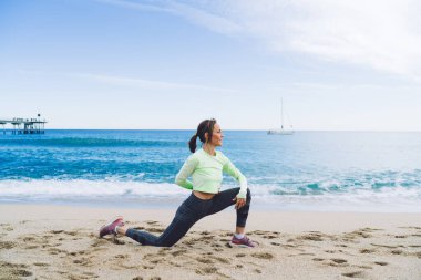 Rahat eşofmanlı esnek kadın vücut geliştirici bacak kaslarını geliştirmek için çömelme hareketleri yapıyor, Kafkasyalı formda kız okyanus kıyısında sabah antrenmanının keyfini çıkarıyor.
