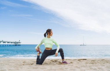 İnce, 40 yaşında, aktif giyimli, siyah saçlı kadın. İskelenin yanındaki kumlu sahilde yoga yaparken destek bacağıyla kıvrılıyor.