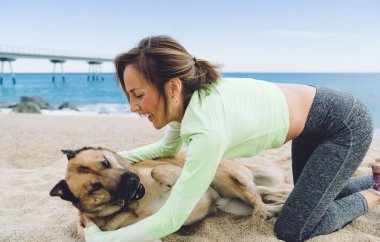 Neşeli zinde bir kadın, deniz kıyısında boş zamanlarını geçirirken, sevimli köpek yavrusu okşayan, aktif eşofmanlı, neşeli bir kadın.