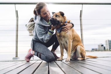 Kırk yaşında kadın, spor kıyafetleri içinde, ahşap köprünün yanında çömelmiş ve köpek okşayan alçak bir açıyla modern korkuluk üzerinde oturuyor.