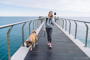 Spor giyim kuşağında tatmin olmuş yetişkin bir bayan köpek tasması takmış, tahta iskelede, akıllı telefondan konuşurken ve gülümserken.