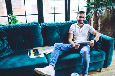 Beyaz tişörtlü, gözlüklü, dişlek gülüşlü rahat koltukta dinlenen ve ofisteki tableti karıştıran etnik sakallı erkek.