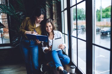 Modern çalışma alanında birlikte çalışırken proje için yeni fikirler yazmak için arkadaş canlısı meslektaşlarının tablet ekranına yaslanan açık defter ve kalemle genç Asyalı kadın.