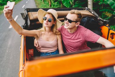Şapkalı ve güneş gözlüklü, gülümseyen sevgilisiyle cep telefonunda selfie çeken kendine güvenen bir kadın yaz gününü yol gezisinde geçiriyor.