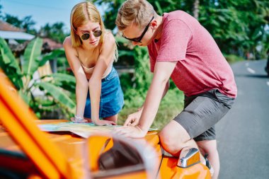 Gündüz vakti güneşli tropikal bir ülkede yaz kıyafetleri ve güneş gözlüğü takan genç çift turuncu araba üzerinde harita çalışıyor.