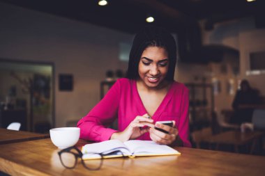 Kafeteryada bir fincan kahveyle oturan genç mutlu siyah kadın ve akıllı telefondan mesajlaşırken planlayıcıyı açtı.