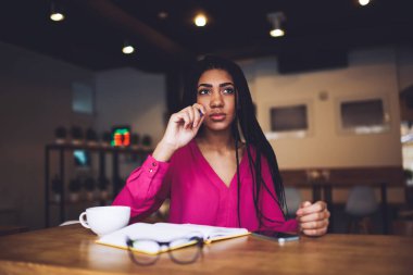 Genç, dalgın, Afro-Amerikalı, gündelik kıyafetleri olan, renkli bir kız öğrenci. Masaya oturuyor, elinde kalemle oturmuş, not defterini açıyor.