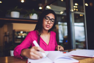 Sade bir kafede sınav hazırlıkları sırasında önemli notlar içeren günlük örgü ve gözlük takan düşük açılı siyahi kız öğrenci.