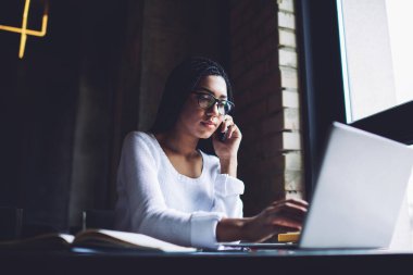 Modern çalışma sahasında dizüstü bilgisayarla yazarken günlük kıyafetiyle cep telefonuyla konuşan genç Afrikalı Amerikalı kadın serbest yazar.