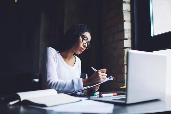 Serious black entrepreneur with black braids in casual clothes taking notes on paper in clipboard while working in dark workspace in daytime