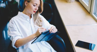 Yukarıdan bakıldığında, beyaz bluzlu, uzun saçlı, masada oturan ve not defterine yazı yazan genç bir kadın görünüyor.