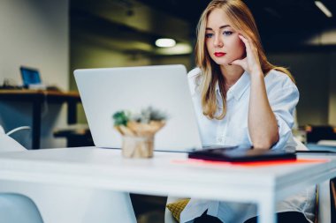 Ciddi bir kadın, modern kafeteryadaki masada uzaktan bilgisayarla çalışırken rahat kıyafetler giyip kırmızı ruj sürüyor.