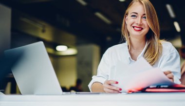 Aşağıdan gülümseyen kadın serbest yazar, netbook kullanıyor ve modern kafede otururken dokümanları kontrol ediyor ve başka yere bakıyor.