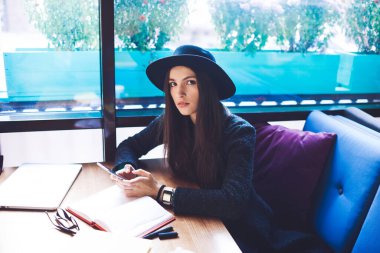 Şık giyinmiş ciddi bir bayan öğrenci elinde açık not defterleriyle masaya oturmuş kameraya bakıyor ve bilgilendirmeleri kontrol etmek için akıllı telefon kullanıyor.