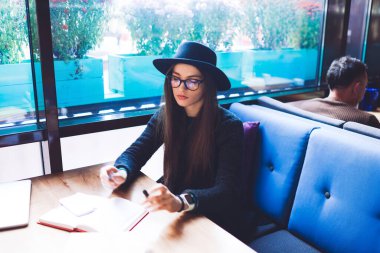 Yukarıdan, şapka ve gözlüklü ciddi bir kadın pencerenin yanındaki masada oturmuş kitap okurken aşağı bakıyor.