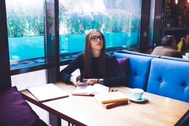 Uzun saçlı, dalgın bir kız öğrenci elinde kahve fincanıyla masaya oturuyor ve cep telefonu kullanırken defteri açıyor.