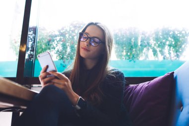 Gözlüklü sakin bir kadın kafeteryadaki yastıklarla kanepede oturuyor ve teneffüste cep telefonu uyarılarını kontrol ederken dinleniyor.