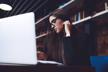 Yeni proje için bilgi toplarken gözlüklü, saçına dokunan ve dizüstü bilgisayar kullanan ciddi bir kadın uzaktan kumanda.